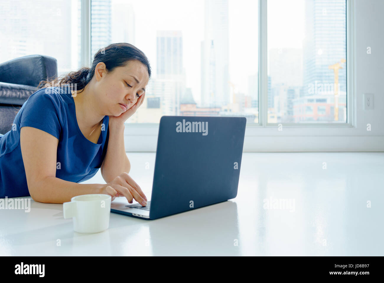 Donna in ambienti chiusi, la posa sul pavimento da window, giorno utilizzando laptop. Asian, capelli lunghi, per adulti Foto Stock
