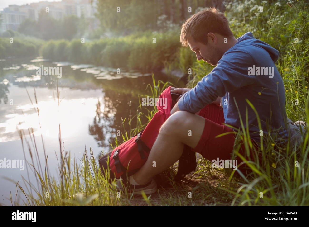 Giovane uomo seduto accanto al fiume con borsa, alla ricerca di qualcosa in borsa e godere nella giornata di sole. Foto Stock