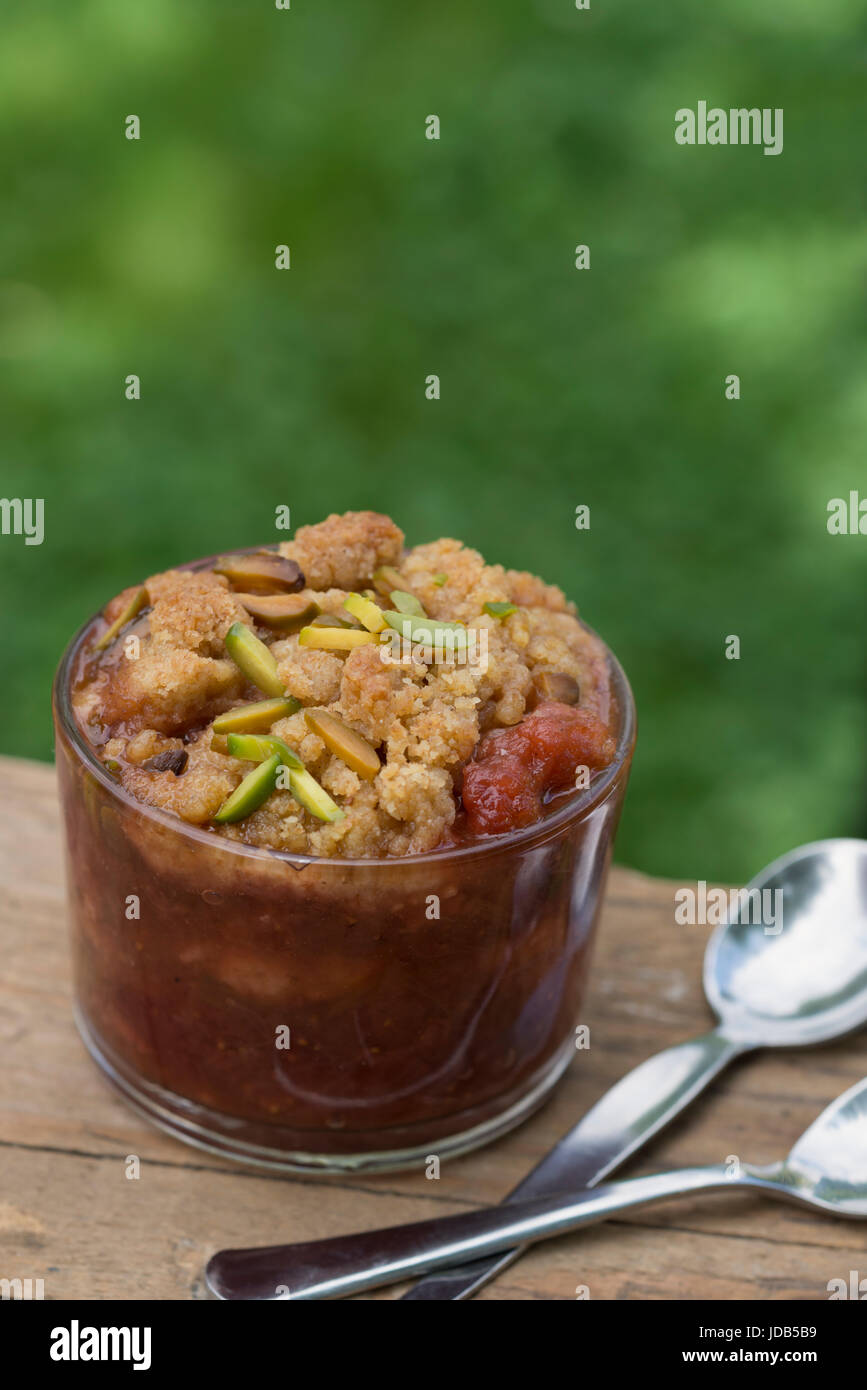 Un bicchiere di fragole e rabarbaro crumble, guarnita con pistacchio, con due cucchiai, su di un tavolo di legno in un giardino verde. Foto Stock