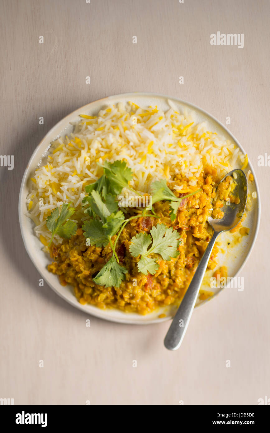 Una piastra di vegano curry di lenticchie con riso basmati, guarnita con foglie di coriandolo fresco, su una luce sullo sfondo di legno con un cucchiaio. Foto Stock