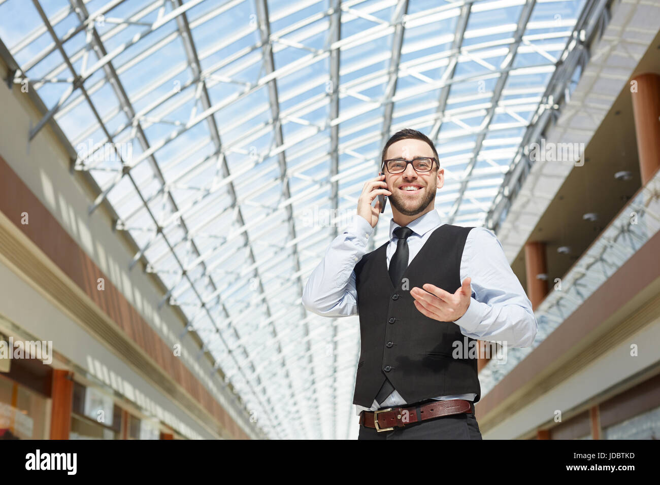 Ritratto di imprenditore moderno che parla al telefono e sorridente in ufficio edificio sotto il tetto di vetro, spazio di copia Foto Stock