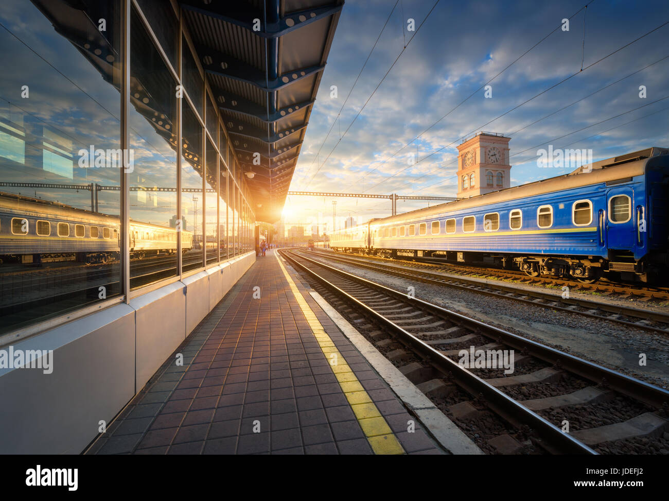 Bella blu treni passeggeri presso la stazione ferroviaria al tramonto. Vista industriale con il moderno treno, ferrovia, piattaforma ferroviaria, edifici e cielo blu Foto Stock