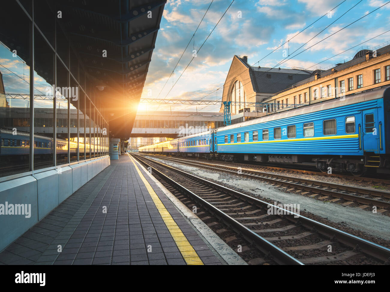 Bella blu treni passeggeri presso la stazione ferroviaria al tramonto. Vista industriale con il moderno treno, ferrovia, piattaforma ferroviaria, edifici e cielo blu Foto Stock