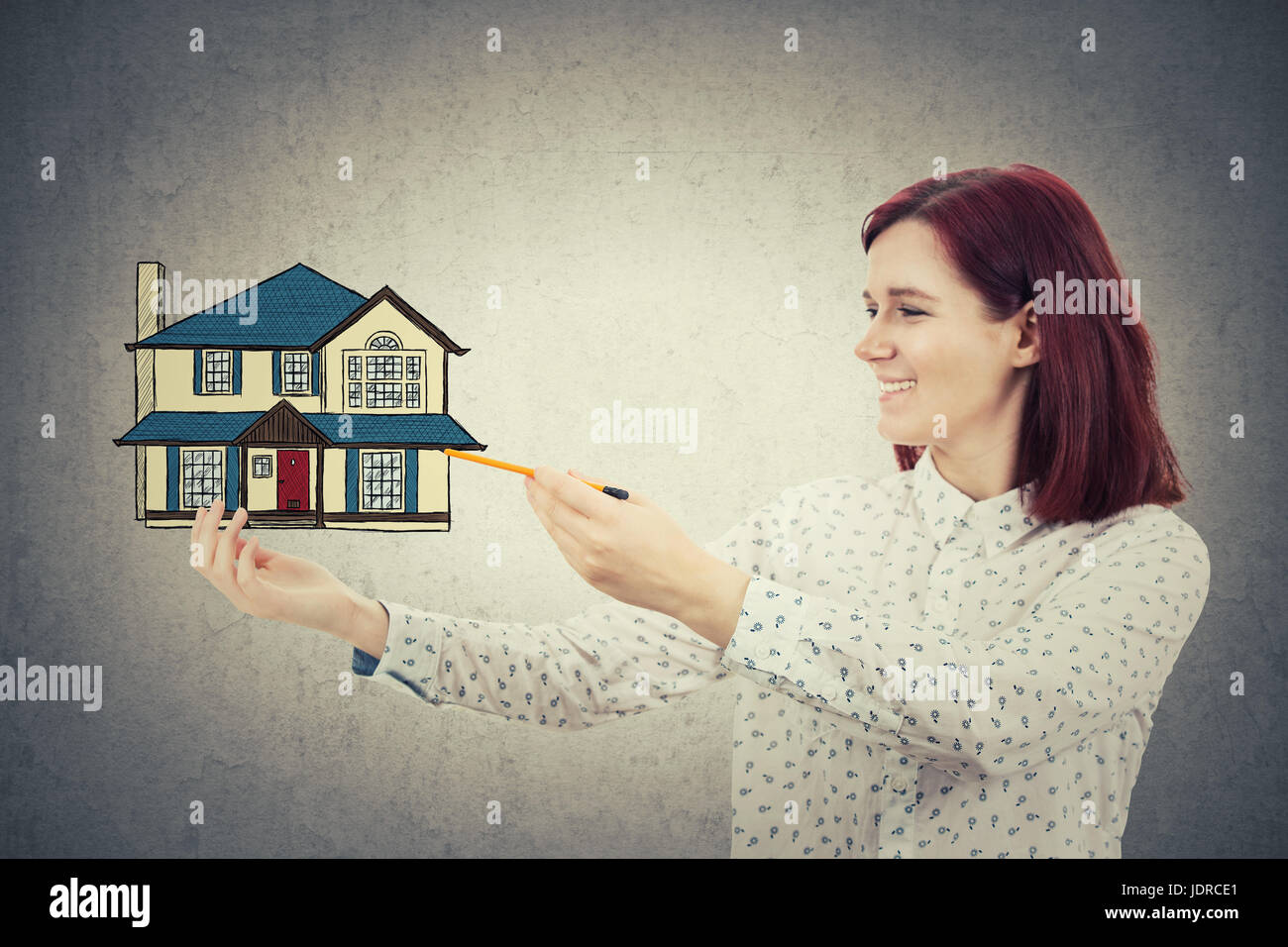 Giovane ragazza sorridente il disegno di una casa di famiglia nella sua mano. Home tutela e assicurazione proprety concetto isolato sul muro grigio Sfondo. Foto Stock