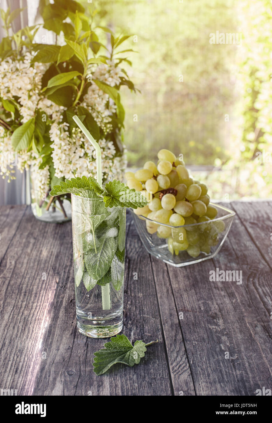 Detox acqua menta, succo d'uva e il ghiaccio in un vetro su uno sfondo di un bouquet di ciliegie e uva in una giornata di sole. Foto Stock