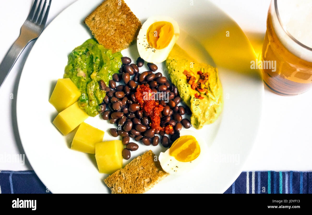 un piatto freddo di cibi misti per l'estate, con una pinta di birra lager fredda: fagioli neri, uova sode, guacamole, hummus, cheddar e toast di grano intero Foto Stock