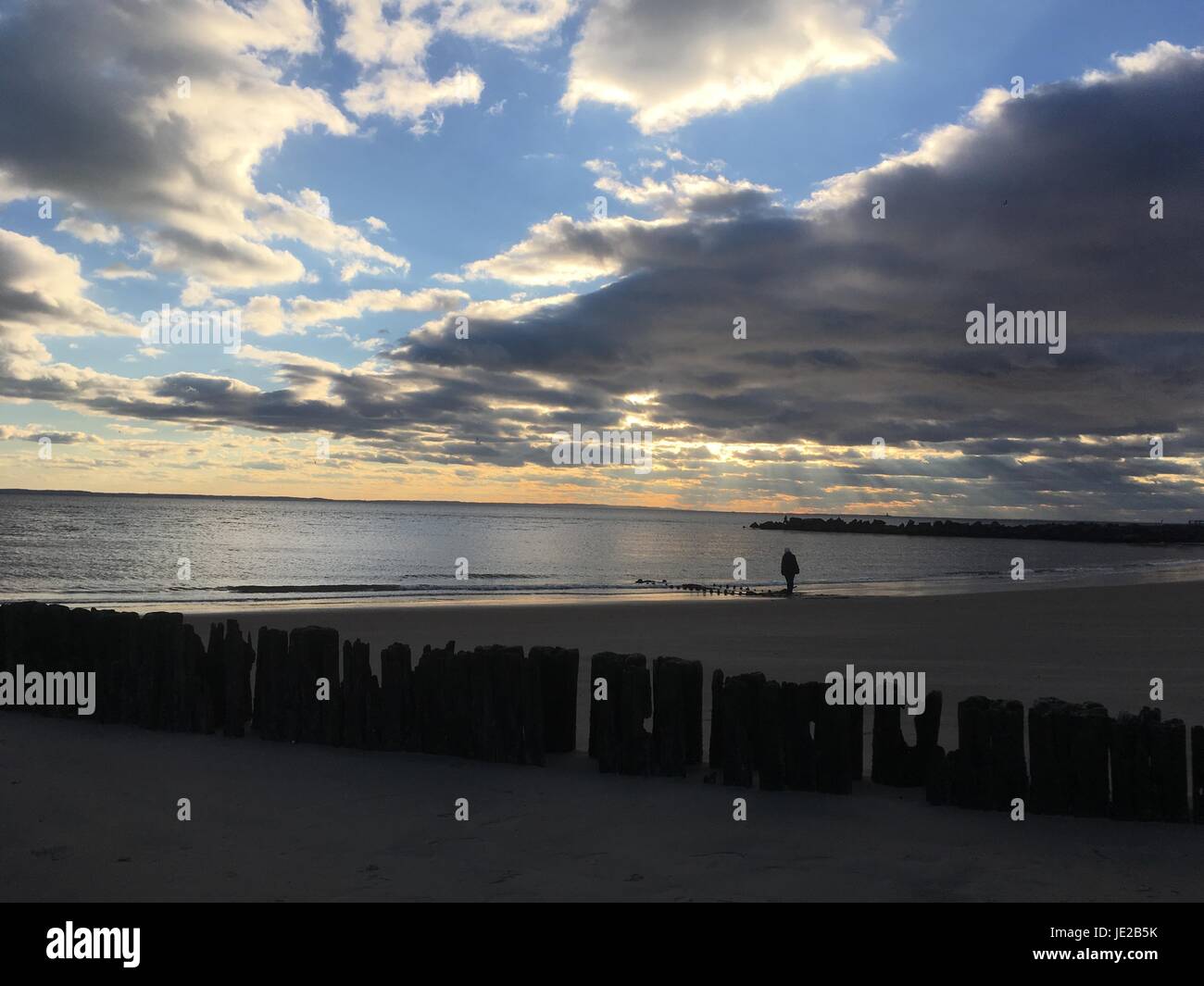 La persona cammina da sola lungo la spiaggia sabbiosa al tramonto. Le nuvole drammatiche si sweep attraverso il cielo. Resti di un molo di legno, una fila di pali di legno intemperie Foto Stock