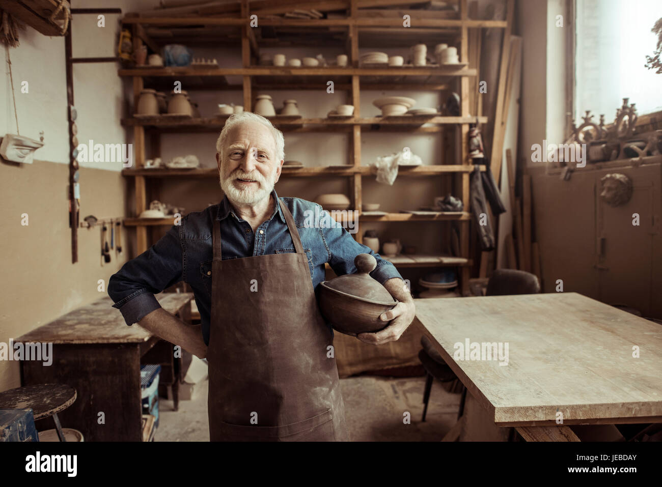 Vista frontale del fiero senior potter nel grembiule in piedi con la ciotola in ceramica in officina Foto Stock