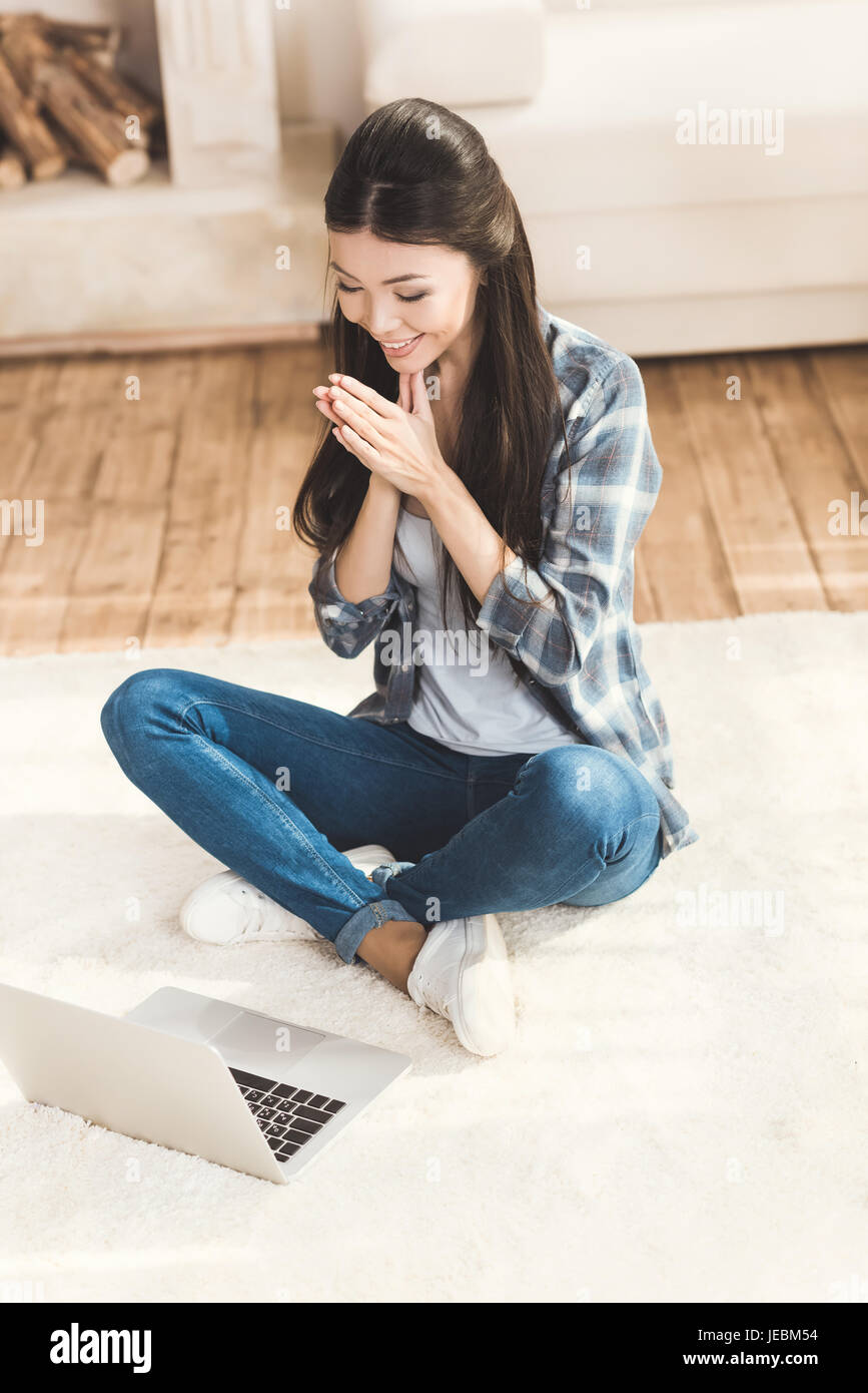 Felice donna seduta sul tappeto e battendo le mani nella parte anteriore del computer portatile Foto Stock
