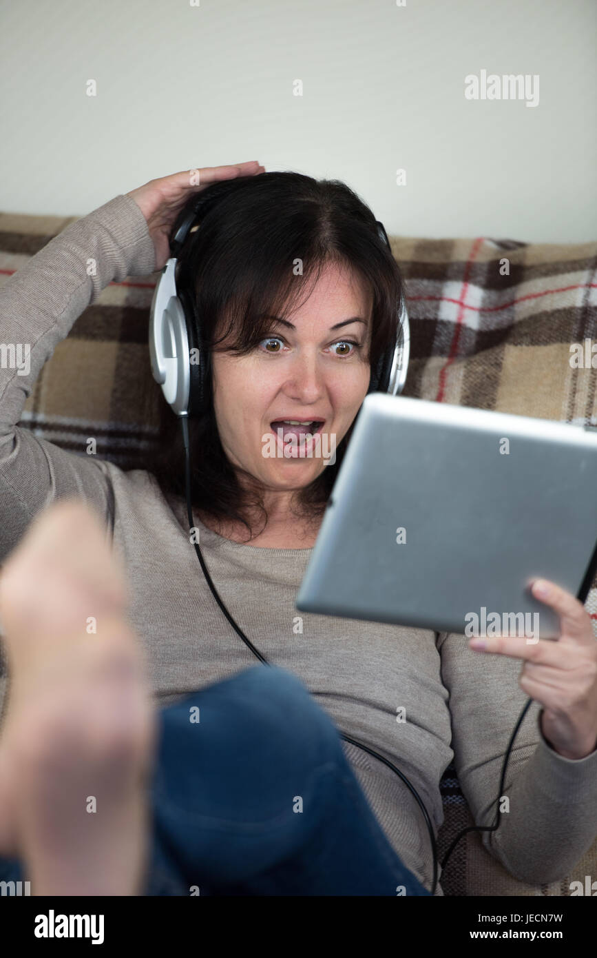 Giovane donna si siede sul divano, rilassante, utilizza un tablet PC per guardare i video, indossa le cuffie Foto Stock Giovane donna si siede sul divano, rilassante, utilizza un tablet PC per guardare i video, indossa le cuffie Foto Stock