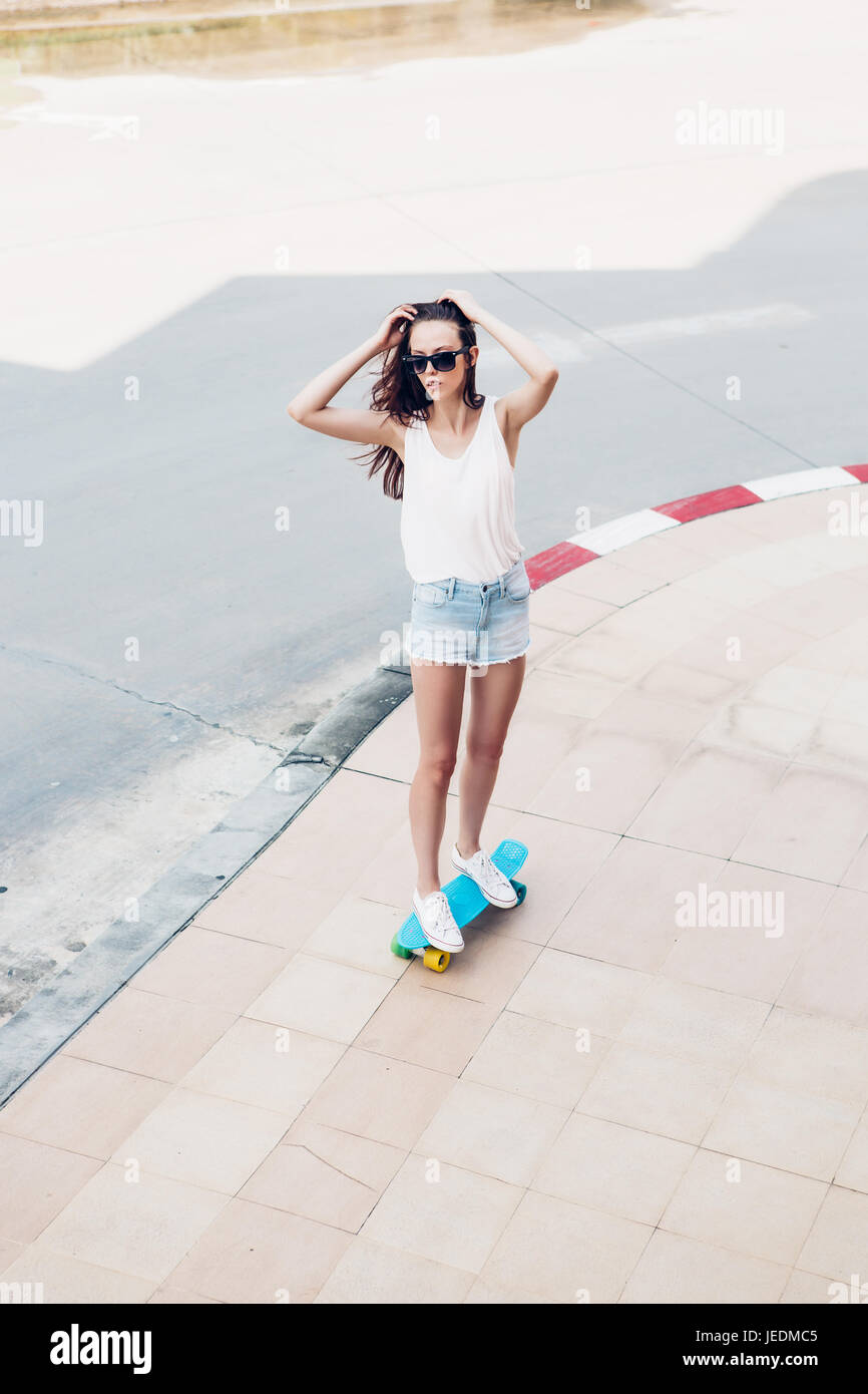 Slim sportivo giovane bella ragazza in bianco t-shirt, jeans brevi cortometraggi e alla moda di occhiali da sole ride blue penny skateboard e correggere i capelli. Scena urbana, Foto Stock