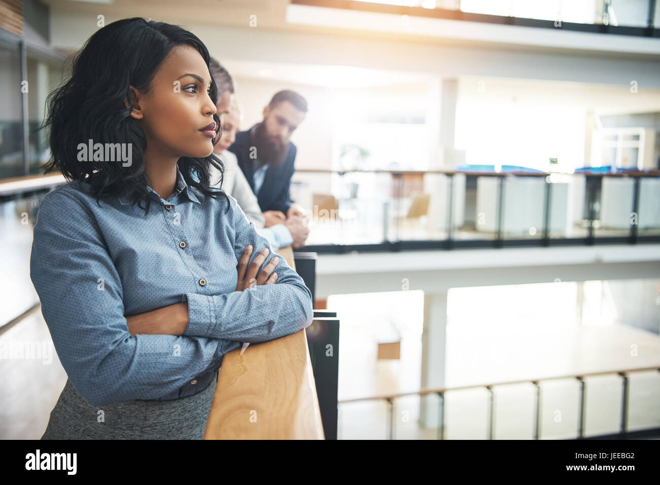 Attraente giovane imprenditrice afro-americano in piedi in ufficio con le braccia incrociate e lo sguardo lontano pensieroso. Foto Stock