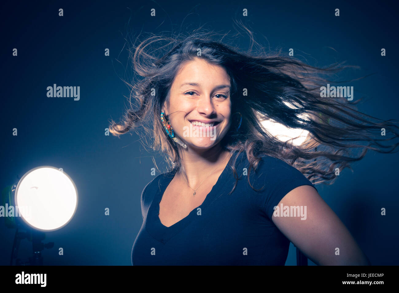 Giovani razza mista donna in tre quarti profilo sorrisi e capovolge i suoi capelli con luci dello studio dietro e uno sfondo scuro Foto Stock