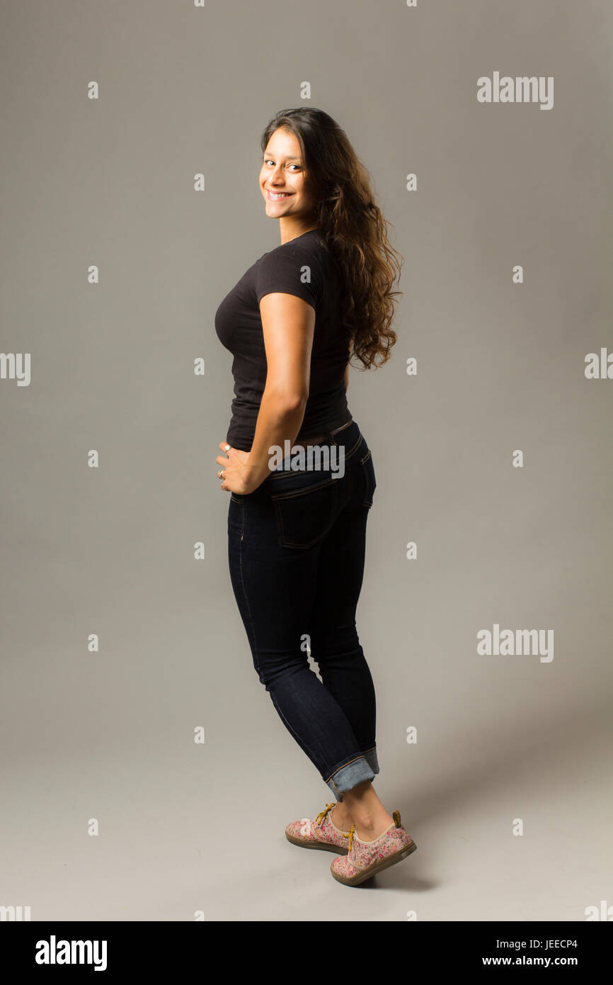 Giovani razza mista donna che indossa un nero di t-shirt e jeans sta guardando sopra la sua spalla mentre sorridente al viewer Foto Stock