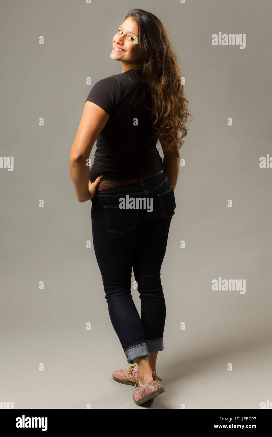 Giovani razza mista donna che indossa un nero di t-shirt e jeans sta guardando sopra la sua spalla mentre sorridente al viewer Foto Stock