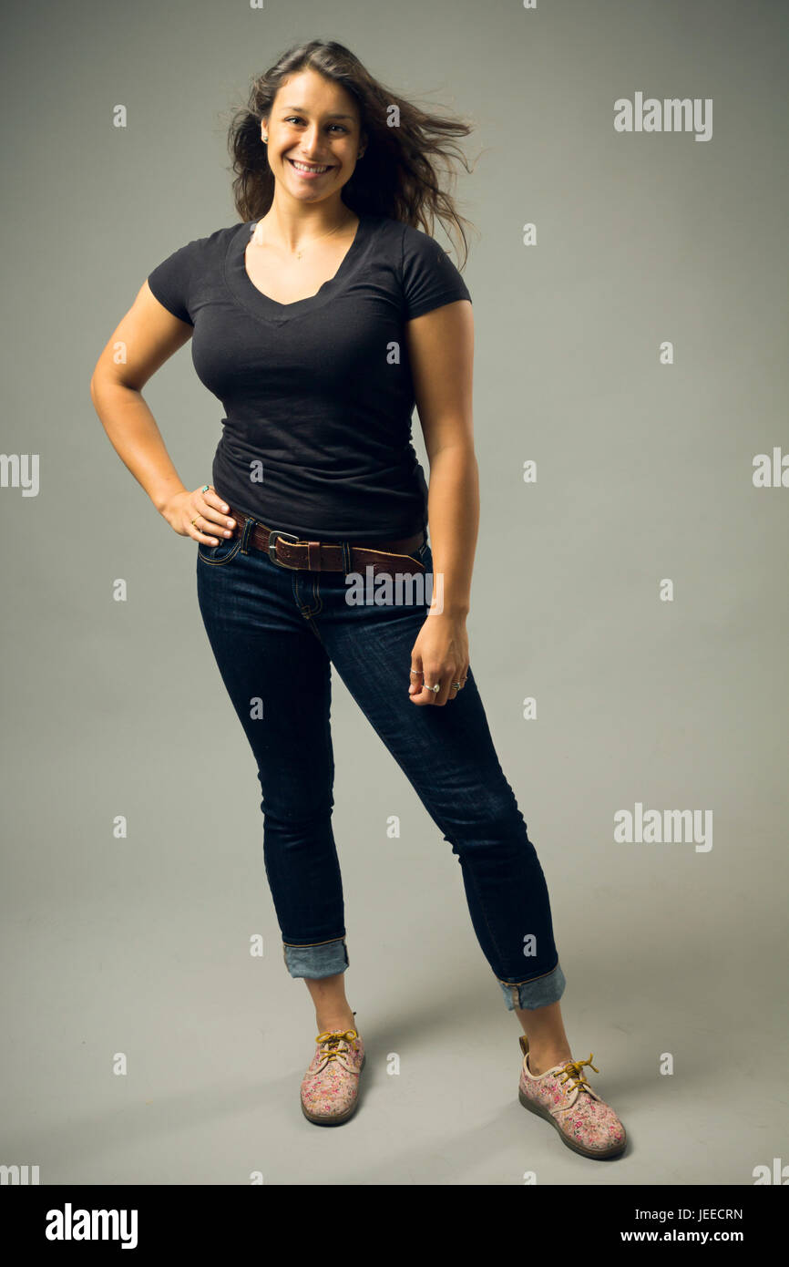 Giovani razza mista donna che indossa un nero di t-shirt e jeans stand con capelli soffiaggio e la mano sul hip mentre sorridente al viewer Foto Stock