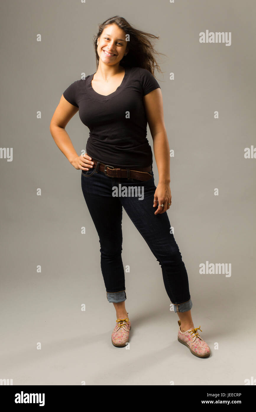 Giovani razza mista donna che indossa un nero di t-shirt e jeans stand con capelli soffiaggio e la mano sul hip mentre sorridente al viewer Foto Stock