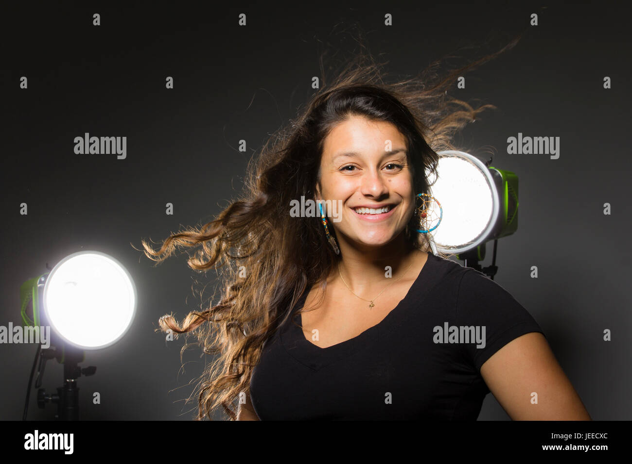 Giovani razza mista donna in tre quarti profilo sorrisi e capovolge i suoi capelli con luci dello studio dietro e uno sfondo scuro Foto Stock