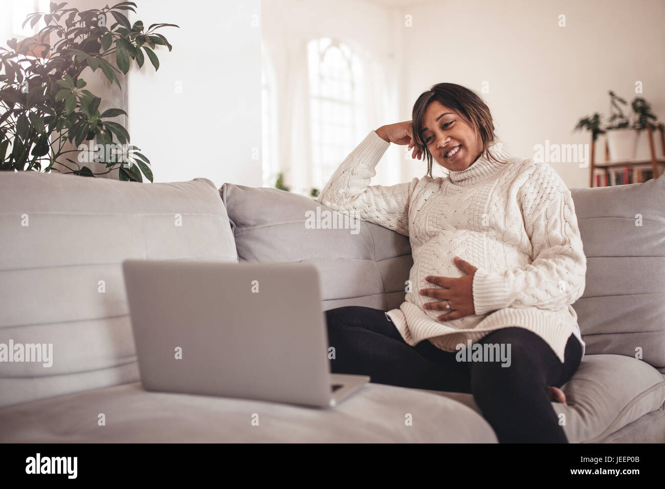 Ritratto di felice giovane donna incinta seduta sul divano e guardando il laptop. La donna in attesa di un bambino seduto sul divano e utilizzo portatile a casa. Foto Stock