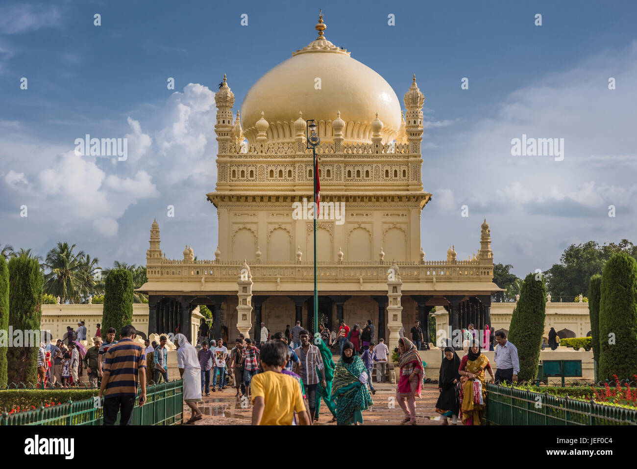 Mysore, India - 26 Ottobre 2013: crema gialla Sultano Tipu mausoleo con cupola sotto pesante cloudscape. Persone che camminano da e per l'edificio. Verde Foto Stock