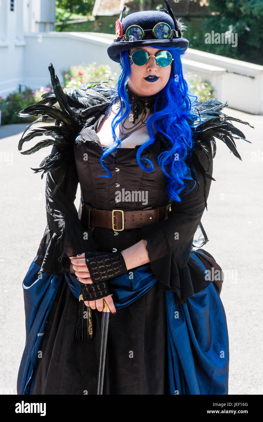 Steampunk donna caucasica, 20s, grandi costruire, in piedi nella luce del sole con il costume nero e il cappello con occhiali, lunghi capelli blu. Foto Stock