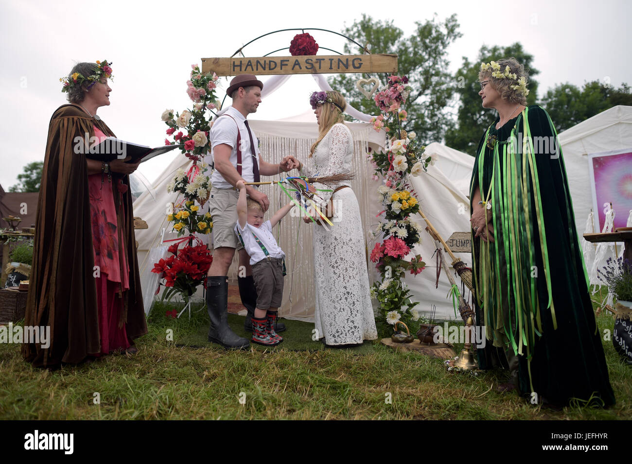 Fi Sexton, 32 e Peter Howe, 38, sia dalla London accompagnato dal figlio Thomas Howe, 2 prendere parte in una Handfasting cerimonia nuziale. La cerimonia eseguita da Glenda proctor (marrone) e Jane Tove (verdi) coinvolte la legatura le loro mani unite con legami, saltando da una scopa e torta di mangiare e di bere birra insieme al festival di Glastonbury, presso l'azienda agricola degna in Somerset. Foto Stock