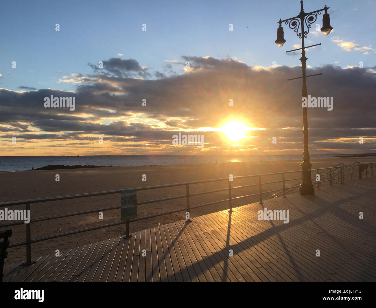 Sole sulla passeggiata di Coney Island, Brooklyn, New York. Lampada da strada in ghisa ornata su molo in legno e ringhiera in metallo. Il sole sorge sulla spiaggia. Foto Stock