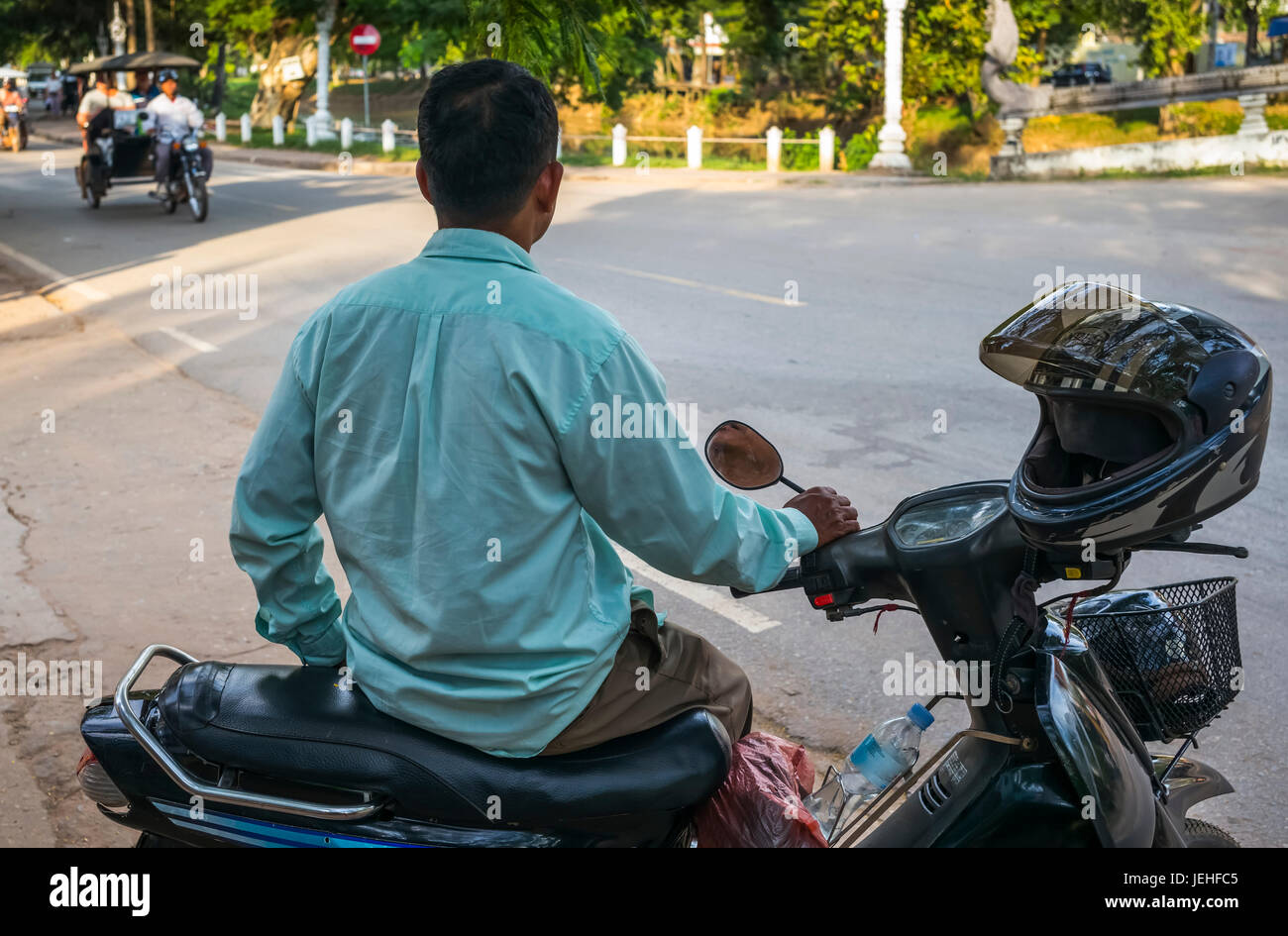Un uomo si siede sulla sua moto a lato della strada come un ciclo rickshaw approcci; Krong Siem Reap, Siem Reap Provincia, Cambogia Foto Stock