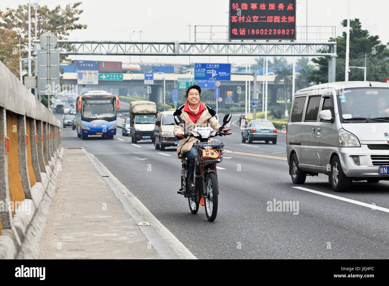 GUANGZHOU-FEB. 21, 2012. Ragazza cinese allegra a cavallo di una bicicletta elettrica su una strada trafficata. Foto Stock