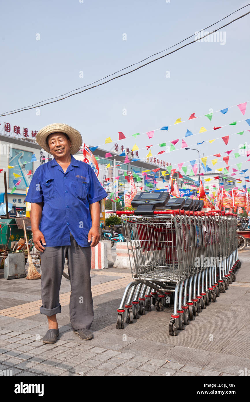 PECHINO-24 LUGLIO 2015. Allegro anziano Wu Mart dipendente con fila caddy. Gli anziani cinesi non possono trovare un lavoro adeguato e spesso accettano uno stipendio basso. Foto Stock