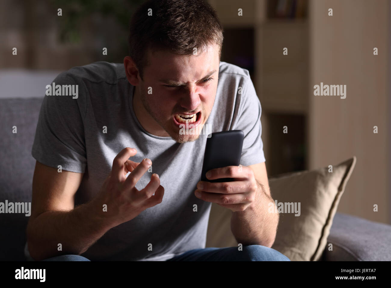 Singola persona arrabbiata con un telefono cellulare seduto su un divano nel soggiorno in una casa interna con uno sfondo scuro Foto Stock