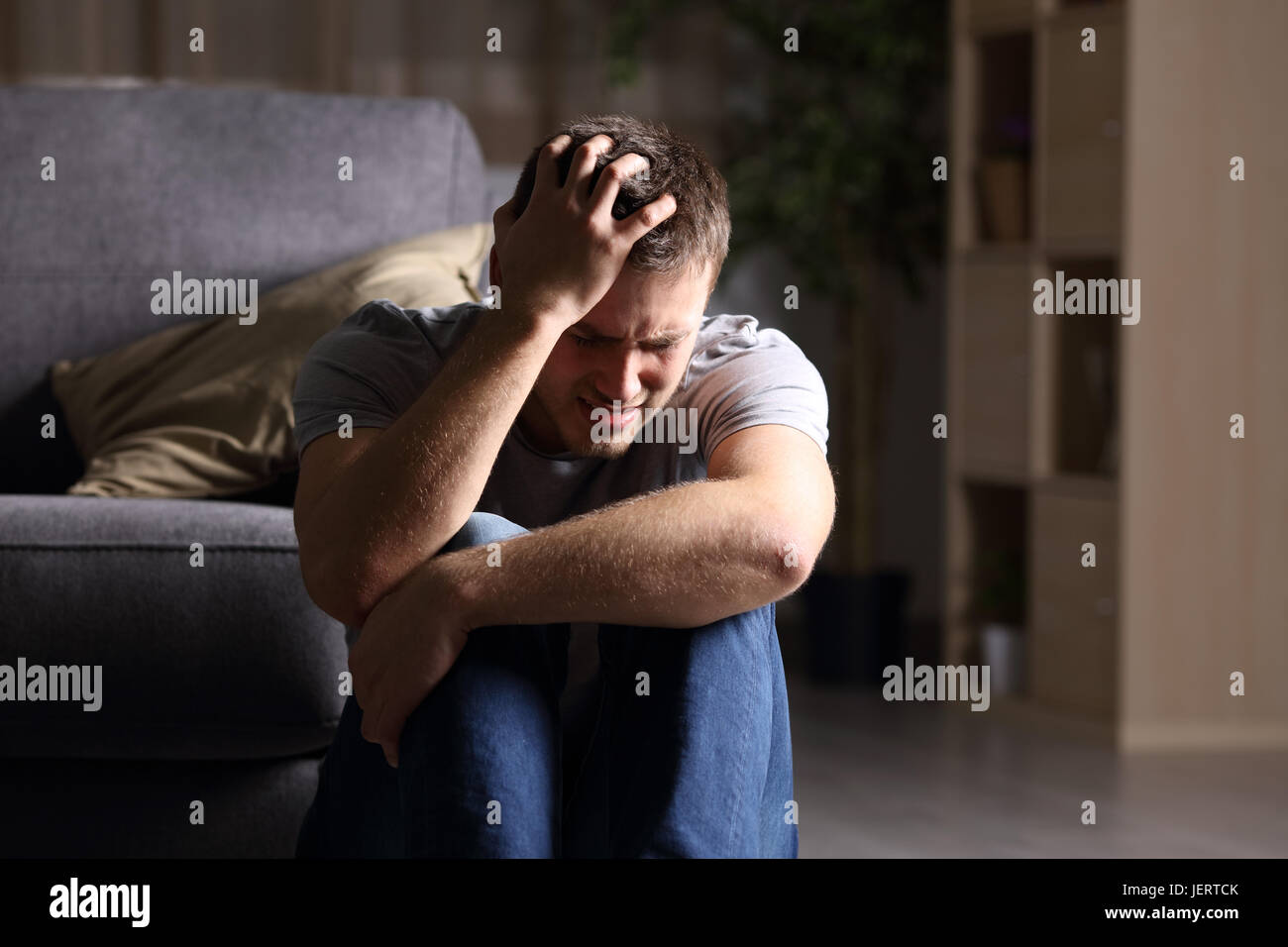 Triste uomo piange seduto sul pavimento del salotto di una casa interna con uno sfondo scuro Foto Stock