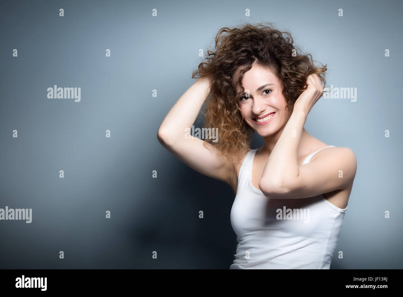 Giovane donna che mantiene i suoi capelli ricci. Bella e sorridente ragazza positiva. Ritratto informale su sfondo grigio. La bellezza naturale e la felicità del concetto. Foto Stock