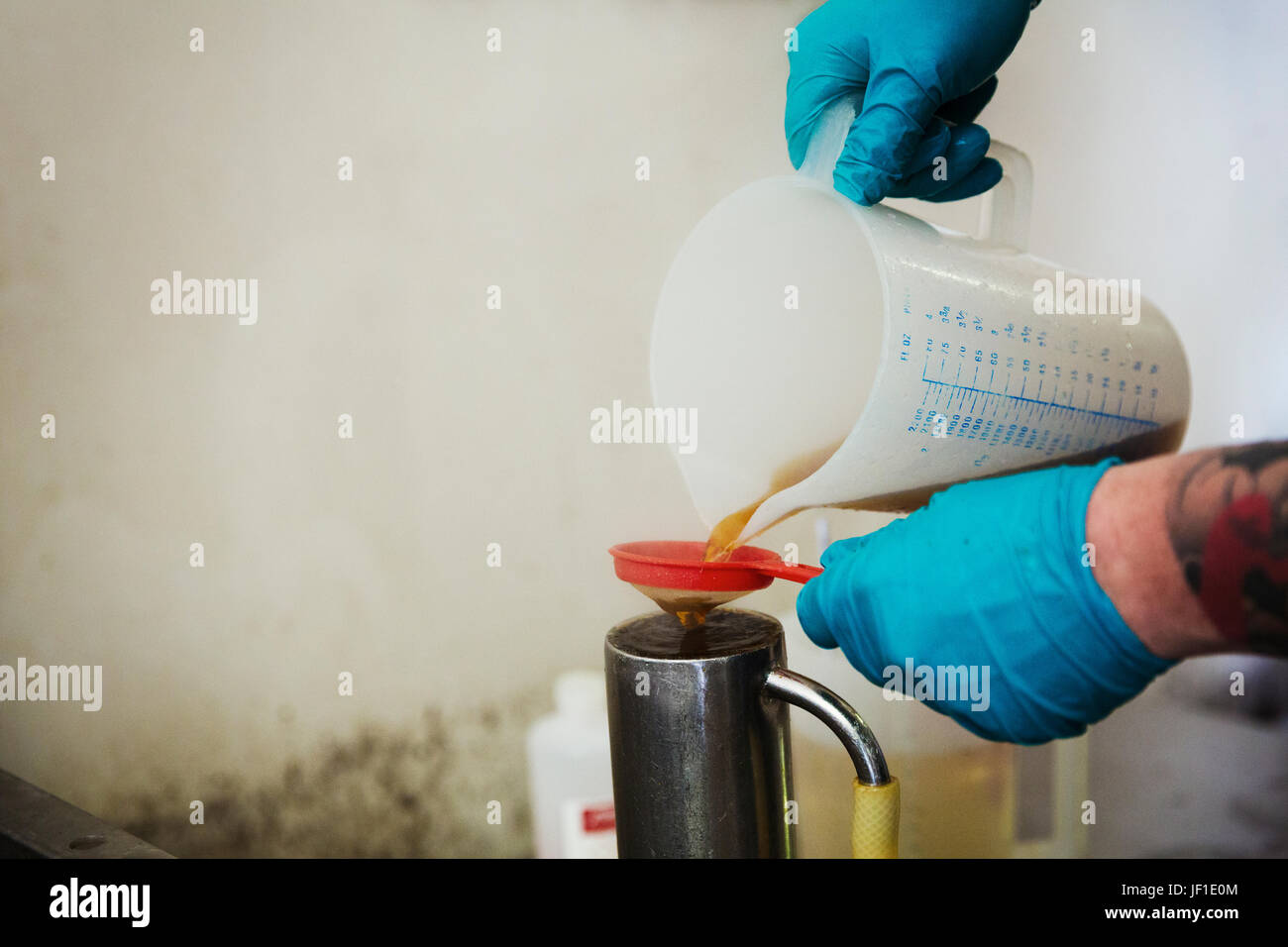 Persona che indossa guanti in lattice blu versando un liquido marrone da una caraffa di plastica attraverso un filtro in una birreria. Foto Stock