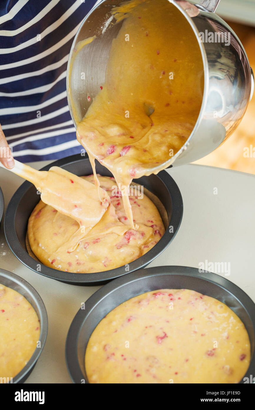 Chiudere fino ad alto angolo di visione della persona che indossa un blu e bianco striata grembiule versando torte da un metallo terrina nel round di teglie. Foto Stock