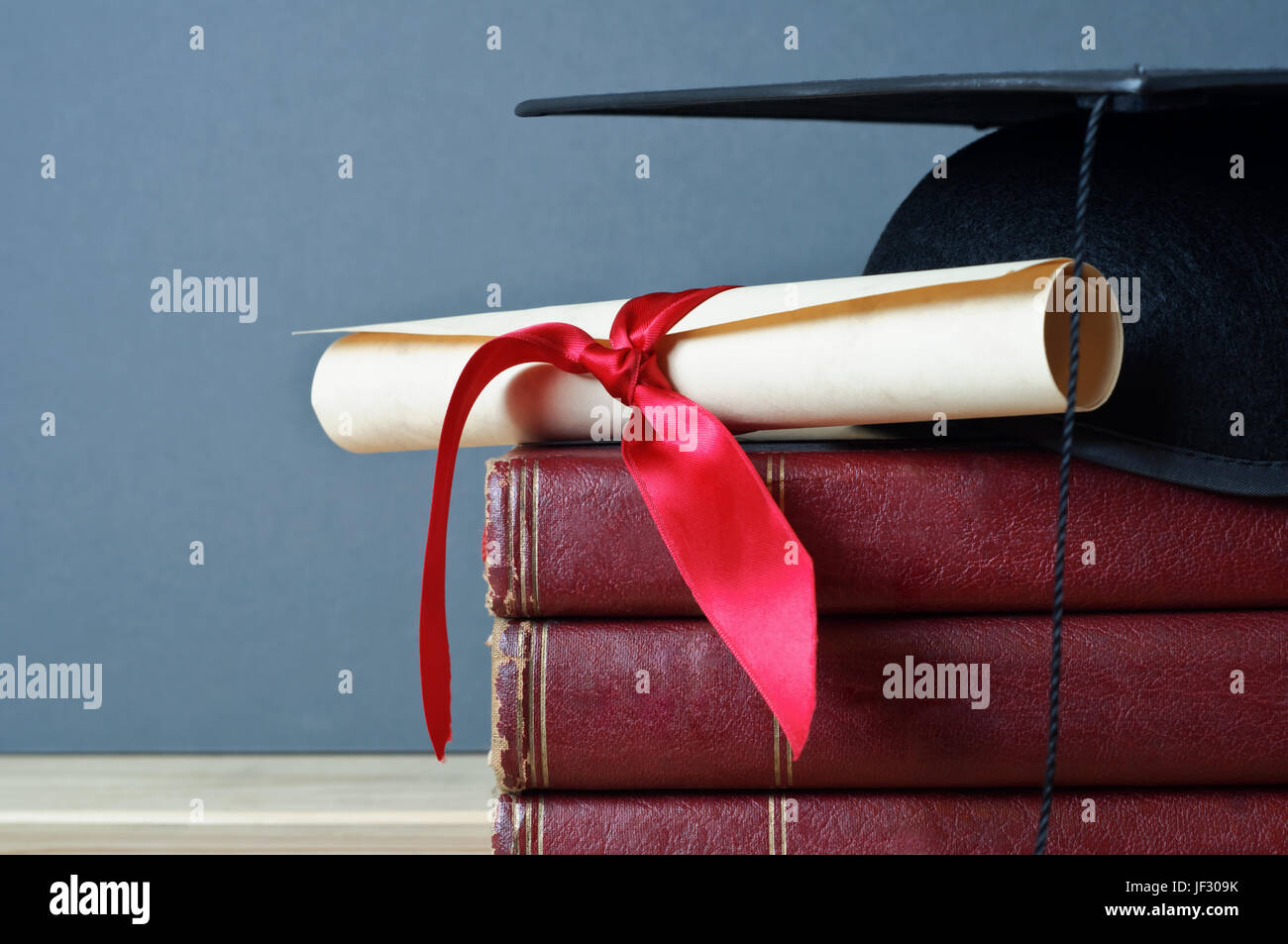 Close up di un mortarboard e graduazione scorrere sulla sommità di una pila di vecchi libri usurati, posto su di un legno chiaro Tavolo con uno sfondo grigio. Foto Stock