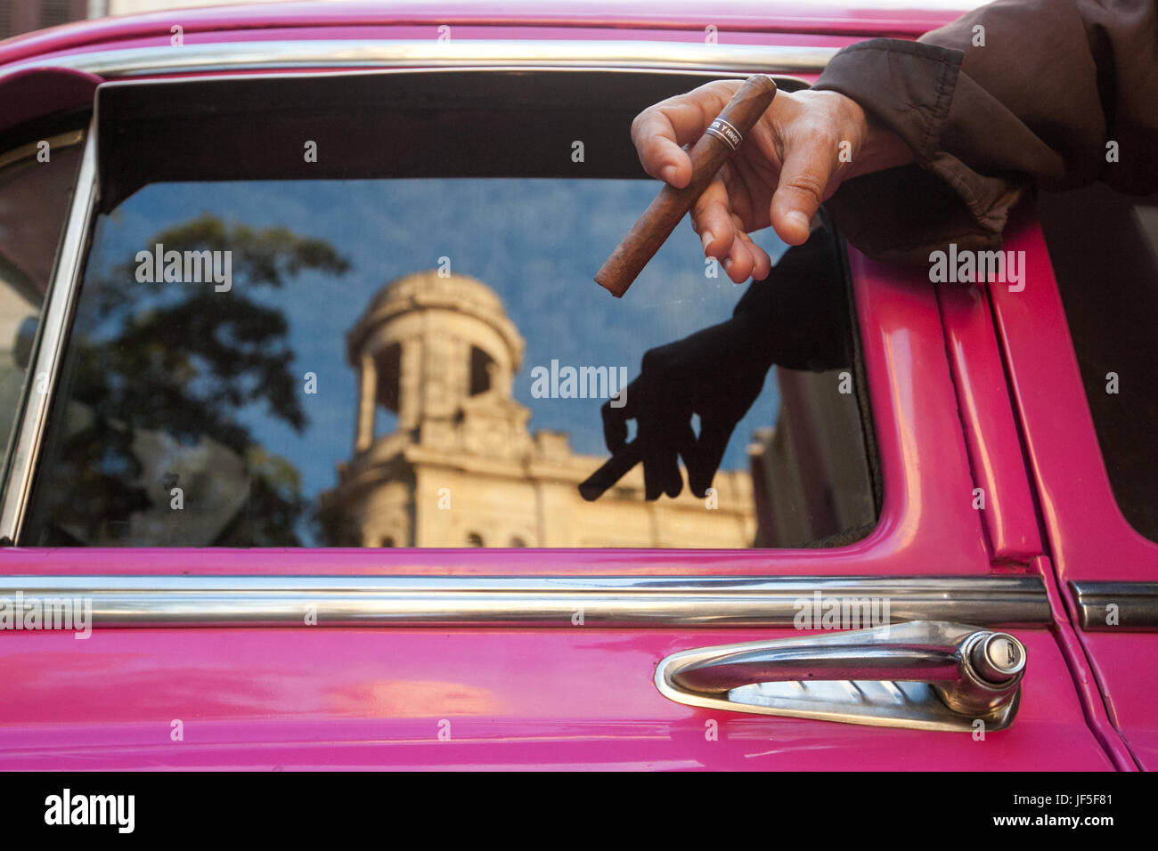 Un uomo detiene un sigaro come architettura cubana è riflessa nella finestra di un colorato classic American car. Foto Stock