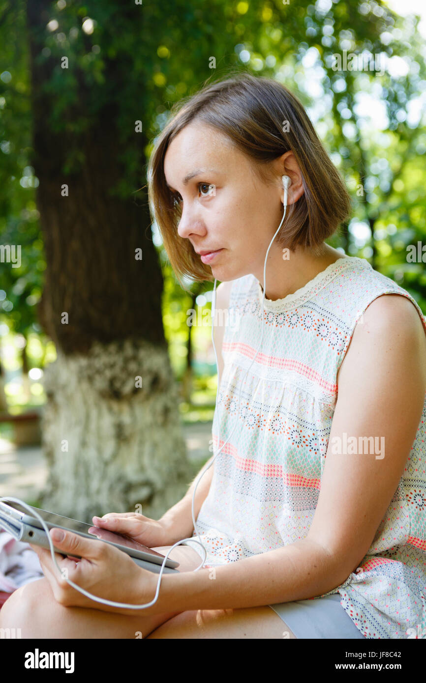 Giovane donna seduta su un fondo di parco e di utilizzare le cuffie con la compressa. Foto Stock