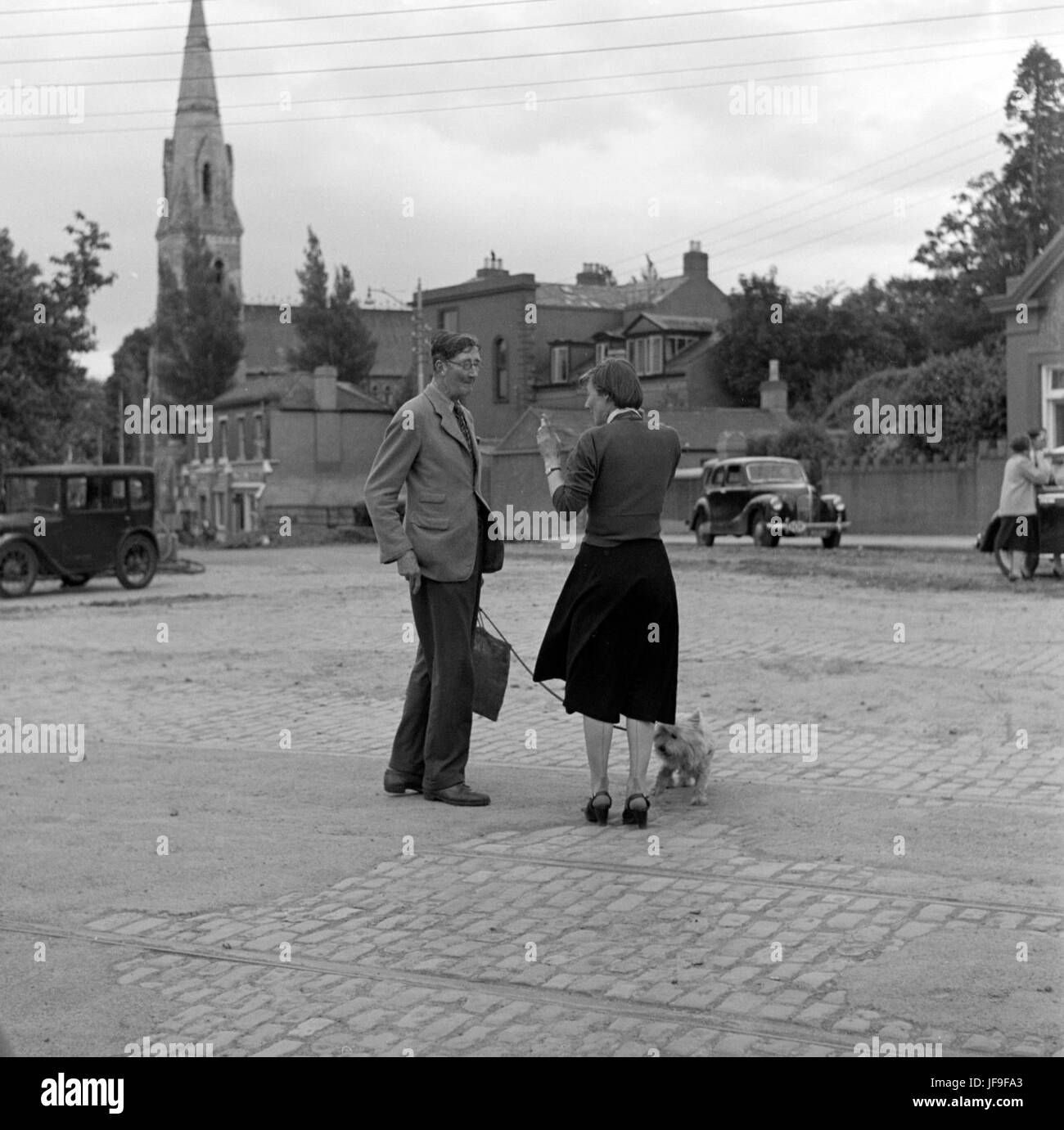 Una fotografia vintage di un uomo e una donna che conversano in una piazza acciottolata, con una chiesa visibile sullo sfondo, che cattura un momento della vita quotidiana. Foto Stock
