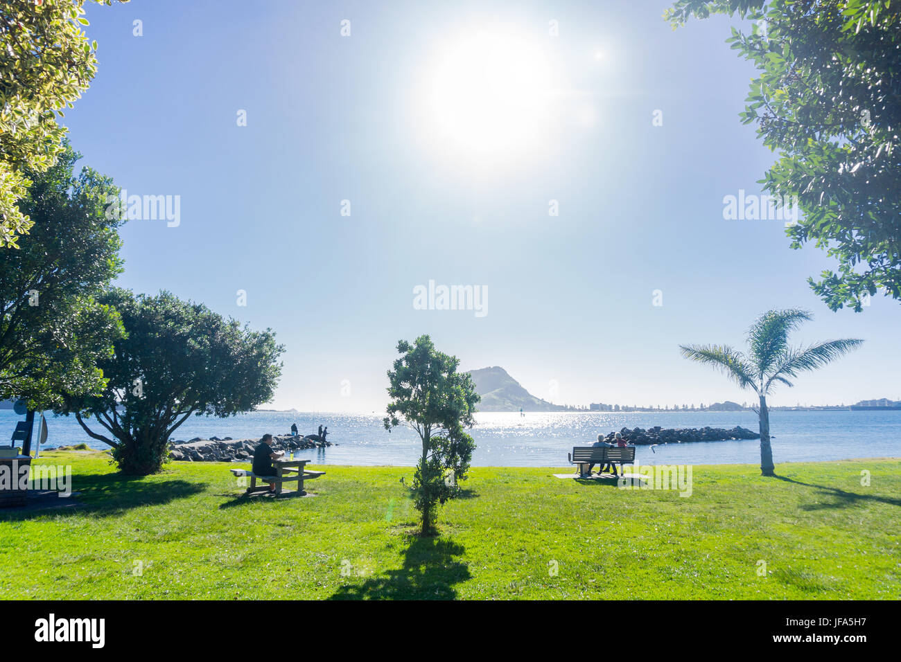 Punto di zolfo scenario, Tauranga vista guardando in sun a Mount Maunganui sull orizzonte lontano con le figure la pesca al largo rock inguine mentre un uomo gode Foto Stock