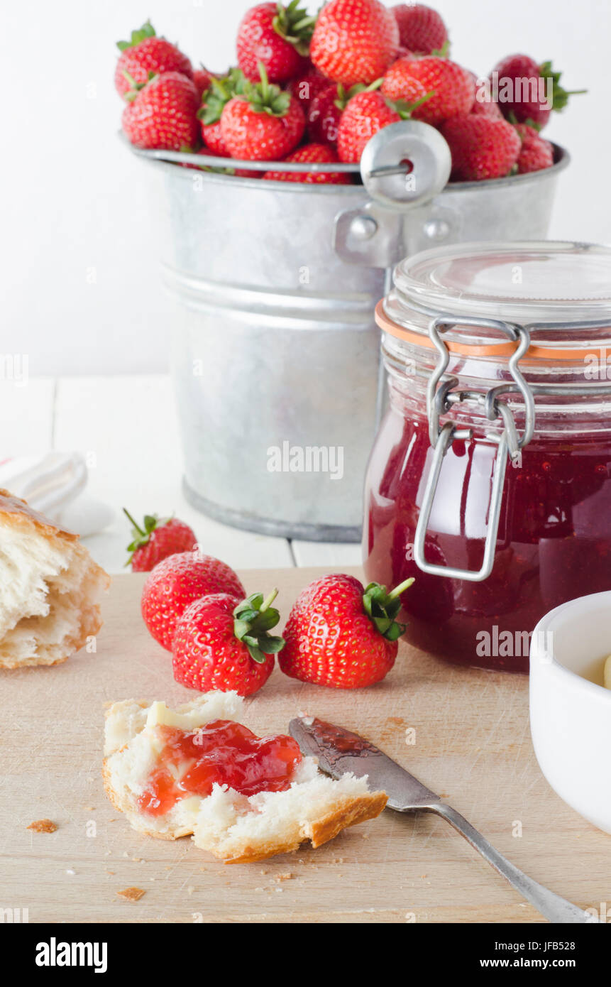Un agriturismo cucina tavola scena di freschi confettura di fragole in vaso di storage con un secchio di stagno di fragole in background e pane e strawber Foto Stock