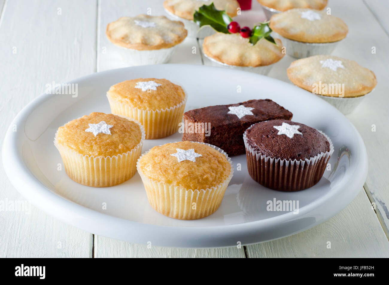 I dolci di natale su una lastra decorata con stelle da setacciato di zucchero a velo, con torte di carne macinata e holly nella messa a fuoco morbida dello sfondo. Steso su un bianco, piano Foto Stock