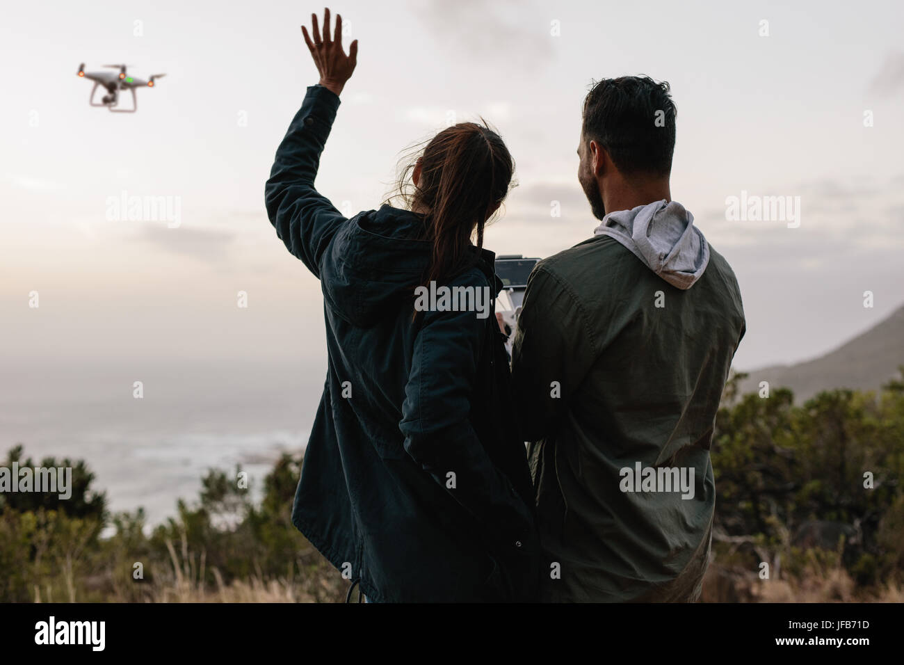 Colpo all'aperto di coppia giovane in piedi insieme in zone rurali e parlando di auto fotografia con un flying drone. Foto Stock