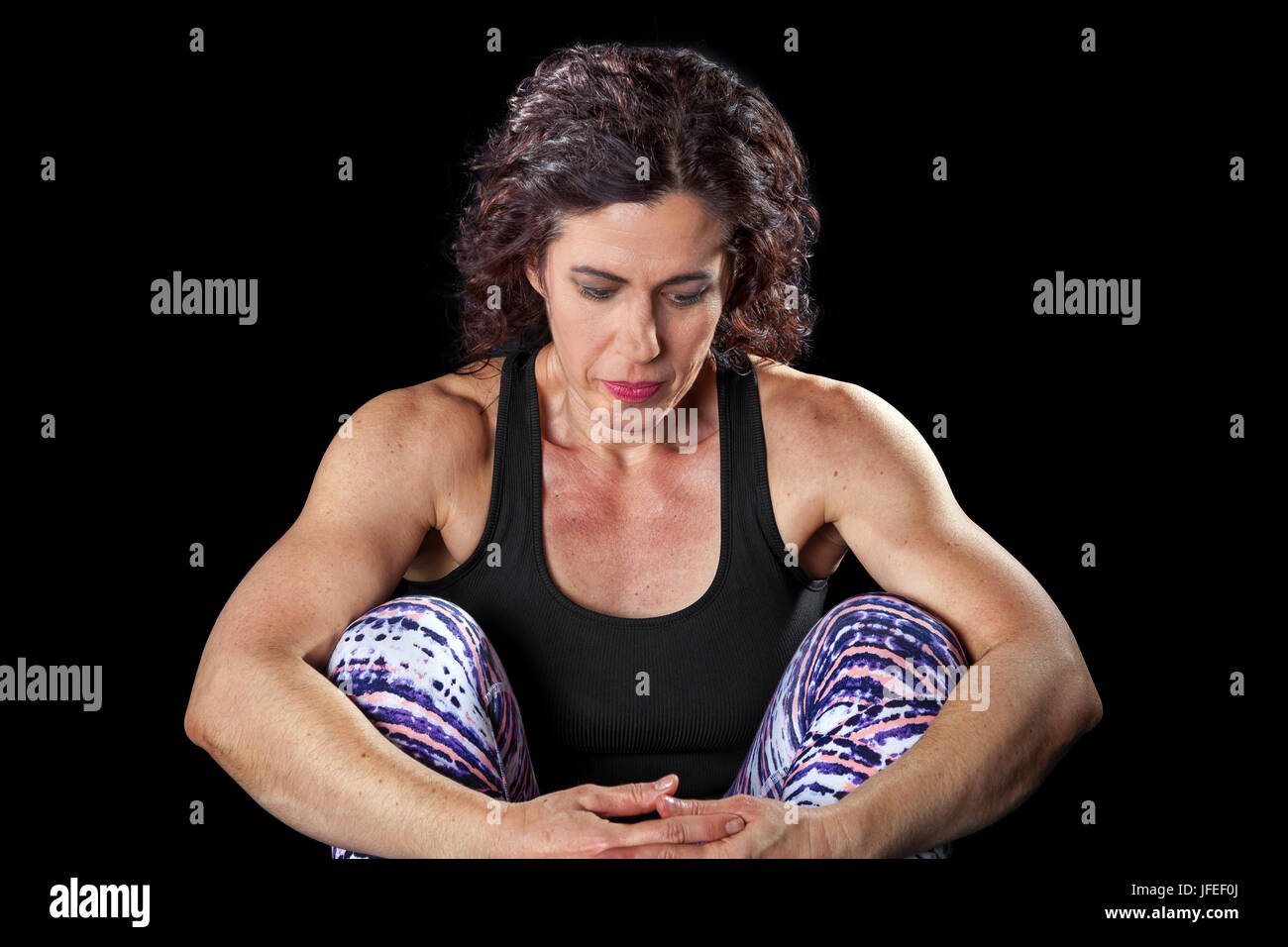 Donna muscolare su sfondo nero si siede con le braccia intorno a lei le ginocchia. Ha un aspetto contemplativo e forse un po' triste o nel profondo del pensiero. Foto Stock
