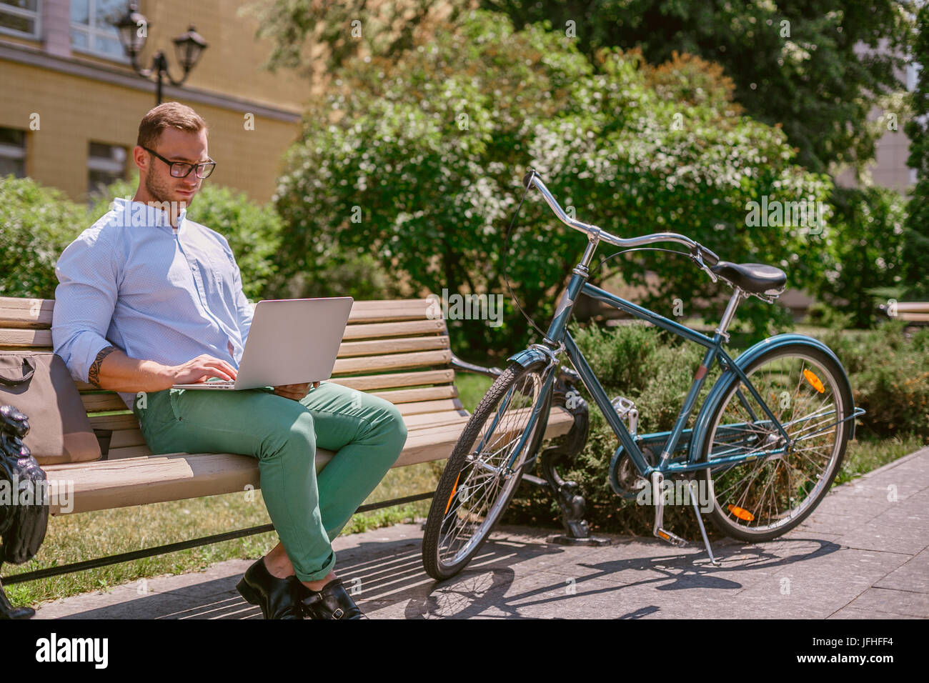 Concentrato imprenditore informale a lavorare con il computer portatile su una pausa caffè. Egli è seduta su una panchina e lavorare al computer portatile, accanto al banco di appoggio di una bicicletta. Foto Stock