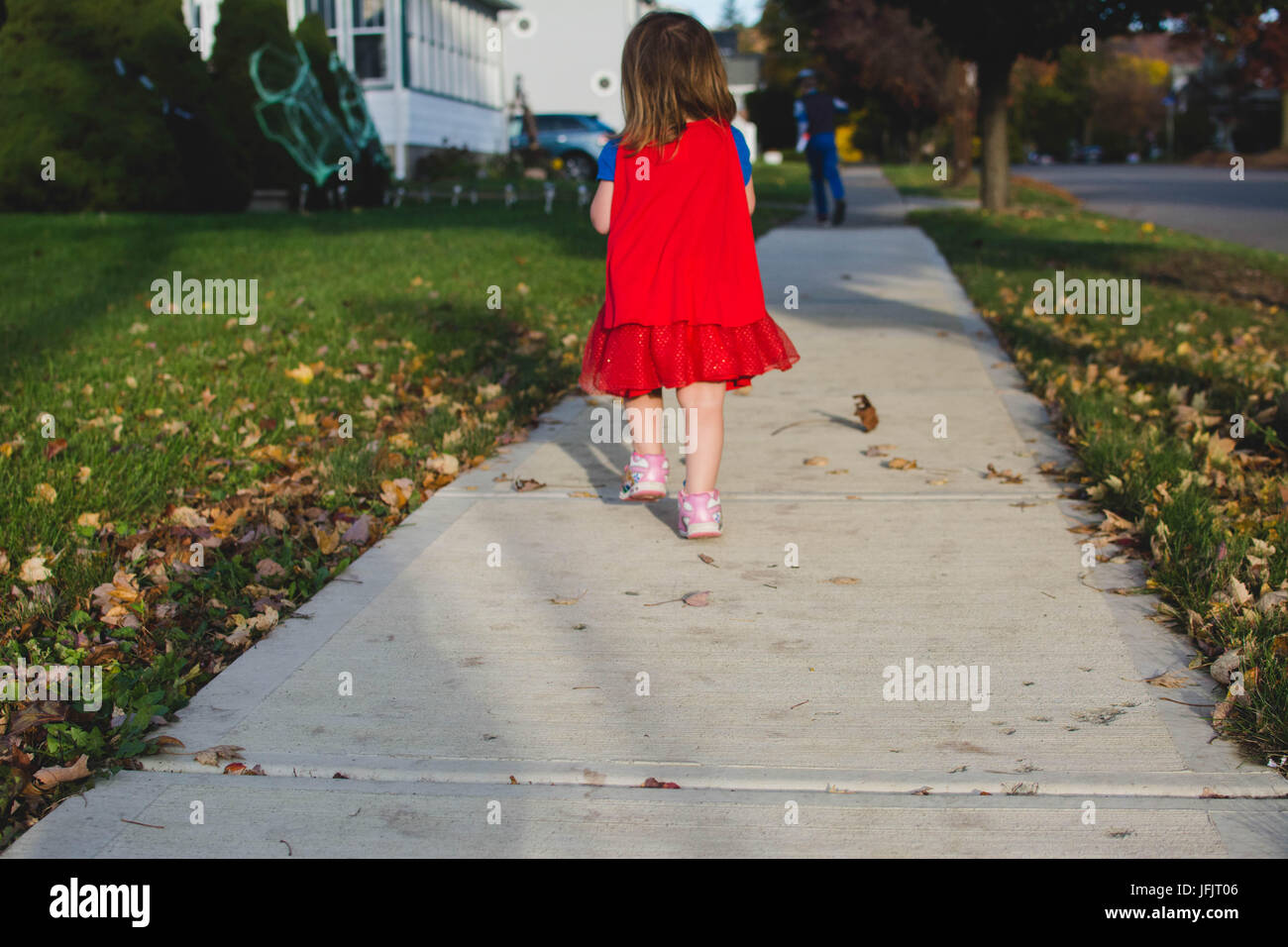 Una giovane ragazza cammina per un marciapiede da dietro al tramonto in autunno che indossa un mantello rosso. Foto Stock
