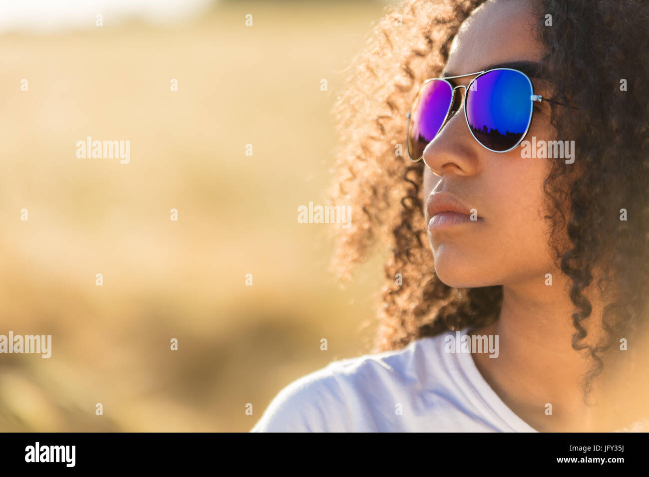 Una bella e premurosa razza mista americano africano ragazza adolescente figlio giovane donna nella luce del sole che indossa blu riflettente aviator occhiali da sole Foto Stock