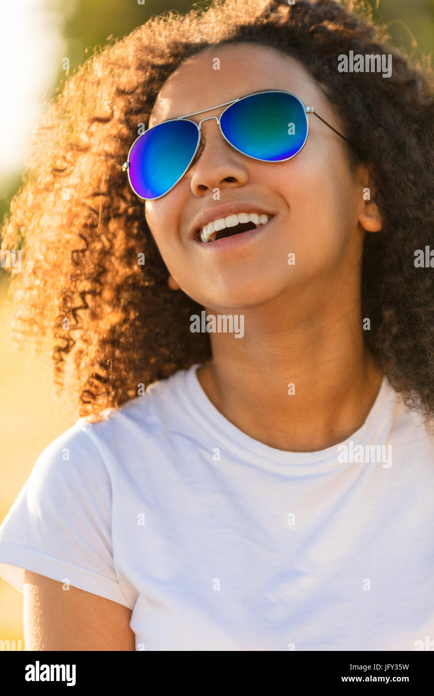 Una bella e felice di razza mista americano africano ragazza adolescente figlio giovane donna nella luce del sole che indossa blu riflettente aviator occhiali da sole e sm Foto Stock