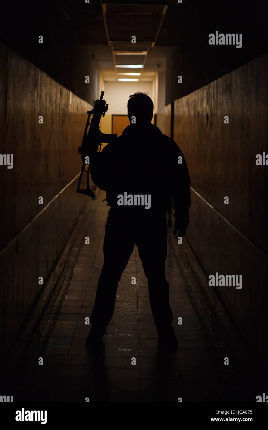 Anti terrorista vestito in camuffamento, in piedi con la pistola nel corridoio scuro illuminato da dietro Foto Stock