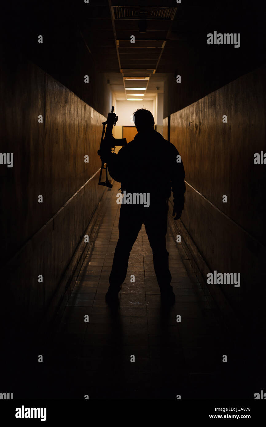 Anti terrorista vestito in camuffamento, in piedi con la pistola nel corridoio scuro illuminato da dietro Foto Stock